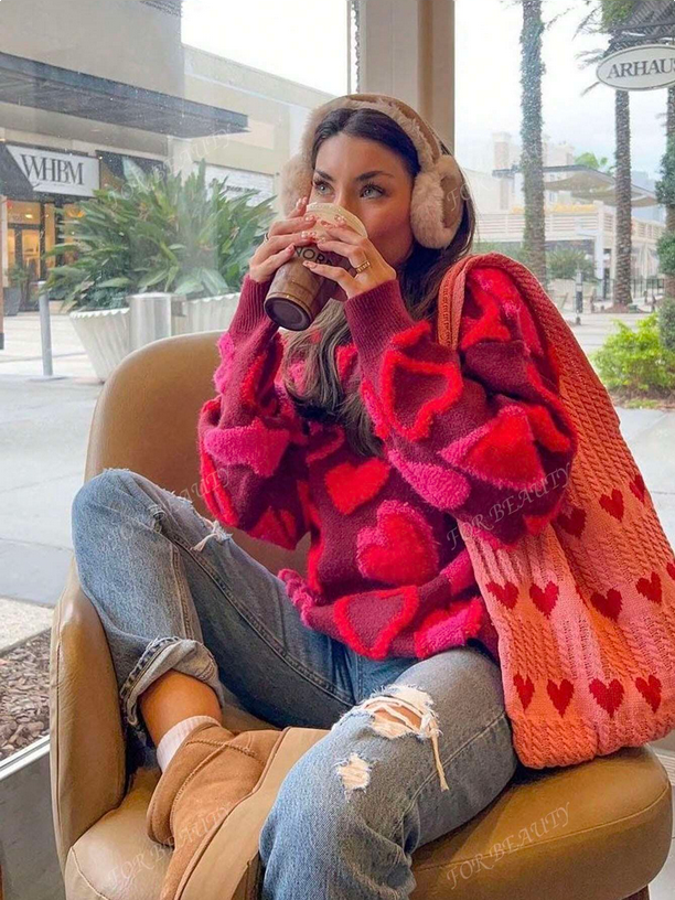 Person wearing a red heart-patterned sweater and ear muffs, sitting on a chair with a drink.