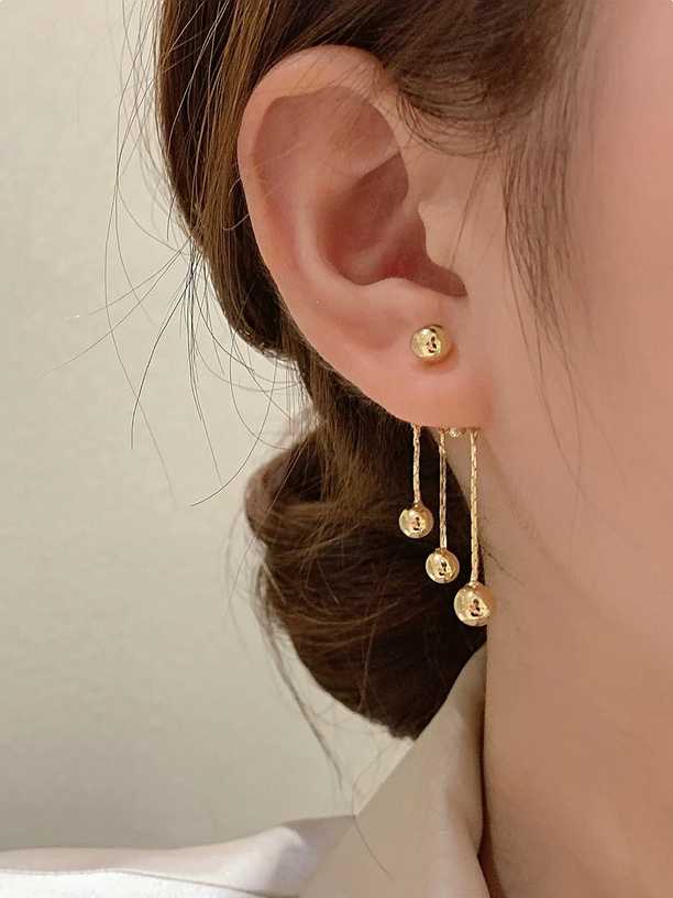 Gold earrings with dangling beads worn by a person against a neutral background
