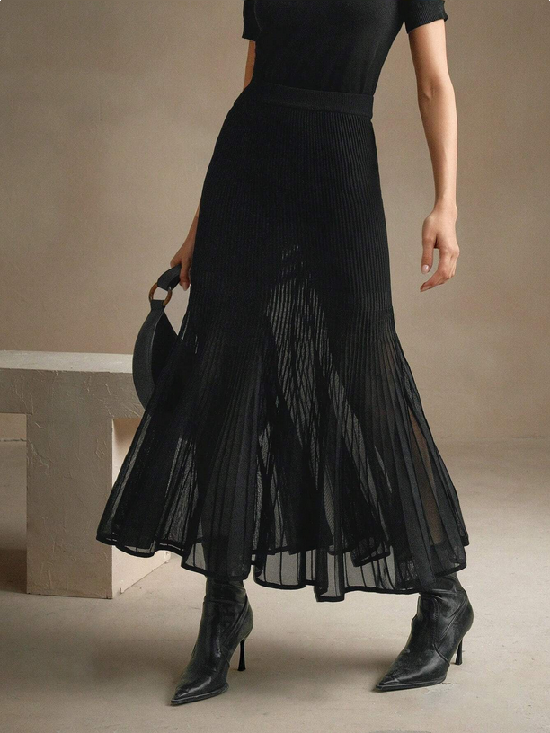 Person wearing a black dress with sheer overlay and black boots in a minimalistic setting