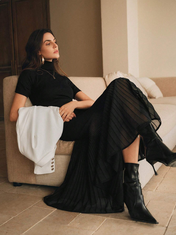 Woman in a black dress and boots sitting on a beige couch in a room with a neutral color palette.