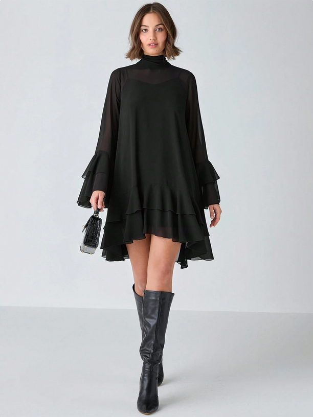 Woman wearing a black dress with sheer sleeves and black knee-high boots on a white background