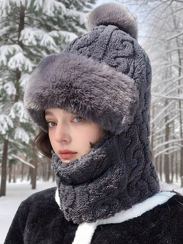 Person wearing a gray knitted hat with a fur trim and scarf in a snowy forest.