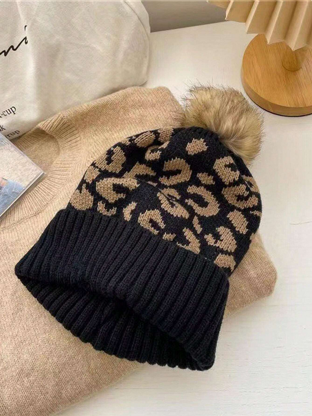 Black knit beanie with leopard print and fur pom-pom on a beige surface.