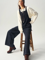 Person wearing denim overalls and a white shirt sitting on a wooden stool against a plain background