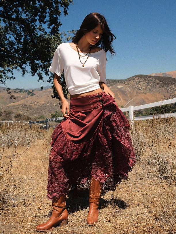 Woman in a white top and burgundy skirt standing in a natural landscape with mountains and trees.