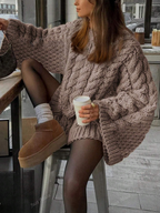 Person wearing a chunky knit sweater and brown shoes, sitting on a stool indoors.